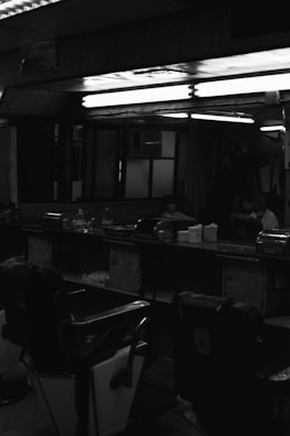 A dimly lit barbershop interior is visible, featuring several empty vintage barber chairs. The space has a classic design with mirrors along the walls, and shelves containing barber supplies and products. The lighting is subdued, creating a shadowy atmosphere within the room.