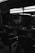 A dimly lit barbershop interior is visible, featuring several empty vintage barber chairs. The space has a classic design with mirrors along the walls, and shelves containing barber supplies and products. The lighting is subdued, creating a shadowy atmosphere within the room.