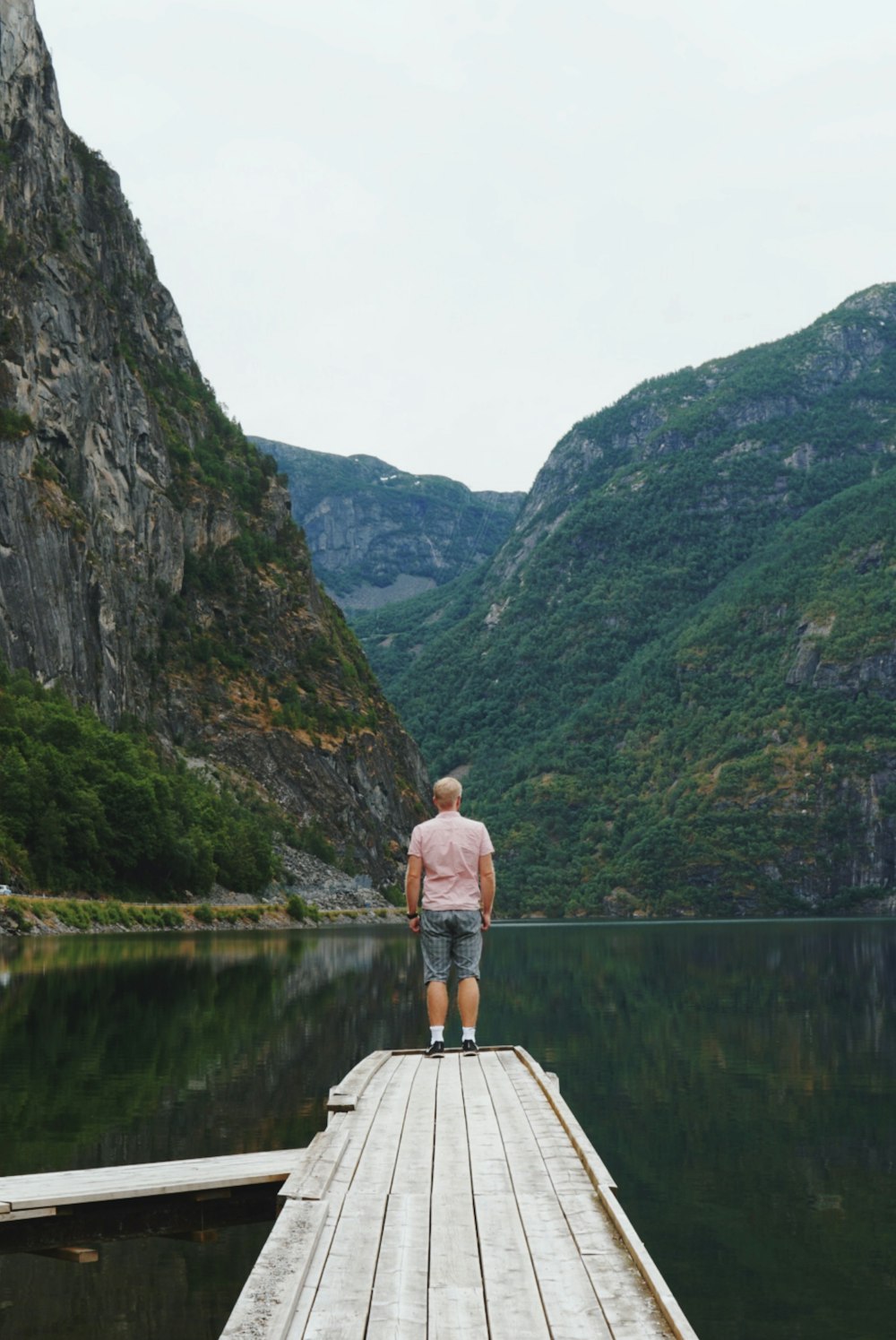 man in brown shorts standing on brown wooden dock near green mountain during daytime