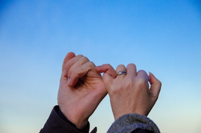 Two hands are linked together by their pinky fingers against a clear blue sky. One hand has a ring on the pinky, and both hands are wearing jackets, suggesting a close bond or promise.