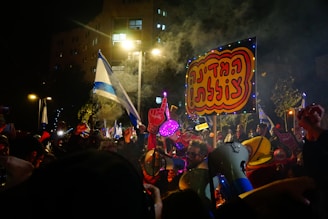 A lively nighttime protest scene with a large crowd of people holding signs and flags. Bright lights illuminate the area, and there's visible smoke in the air. One prominent sign has bold, colorful lettering, and several people are holding megaphones and foam hands.