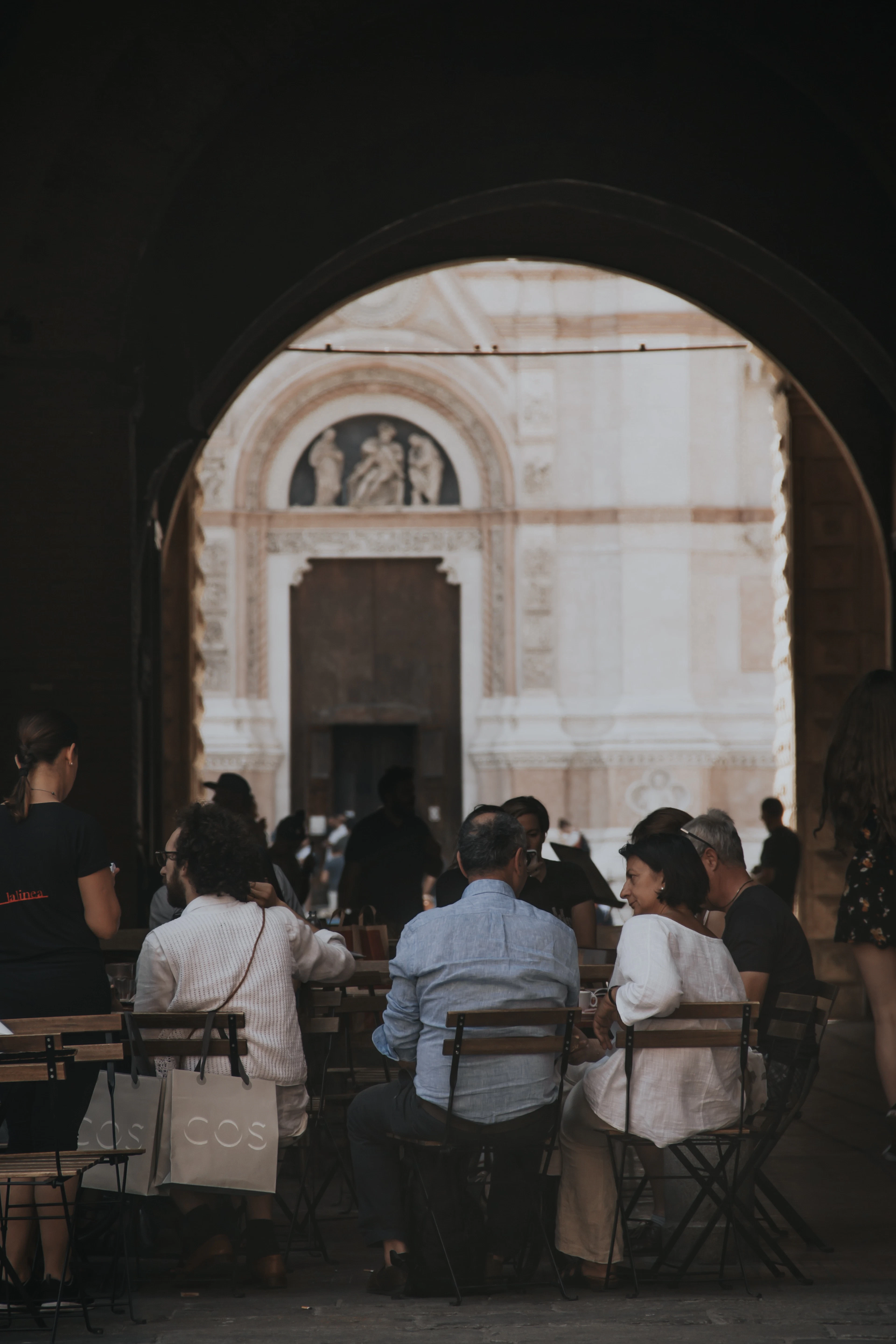 Diners at outdoor tables beneath a shadowed stone portico, framing a sunlit Romanesque church facade beyond.