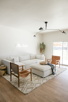 A modern living room featuring a large white sectional sofa with several cushions, accompanied by two leather armchairs with wooden frames. In front of the sofa is a large upholstered ottoman. A soft, patterned area rug covers the wooden floor, and a minimalist, black metal ceiling light fixture hangs above. A potted plant with large leaves is positioned in the corner near a window that allows natural light to flood the room. A small round wooden side table sits next to the sofa with a few items on top.