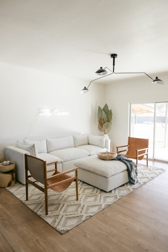 A modern living room featuring a large white sectional sofa with several cushions, accompanied by two leather armchairs with wooden frames. In front of the sofa is a large upholstered ottoman. A soft, patterned area rug covers the wooden floor, and a minimalist, black metal ceiling light fixture hangs above. A potted plant with large leaves is positioned in the corner near a window that allows natural light to flood the room. A small round wooden side table sits next to the sofa with a few items on top.