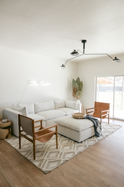 A modern living room featuring a large white sectional sofa with several cushions, accompanied by two leather armchairs with wooden frames. In front of the sofa is a large upholstered ottoman. A soft, patterned area rug covers the wooden floor, and a minimalist, black metal ceiling light fixture hangs above. A potted plant with large leaves is positioned in the corner near a window that allows natural light to flood the room. A small round wooden side table sits next to the sofa with a few items on top.