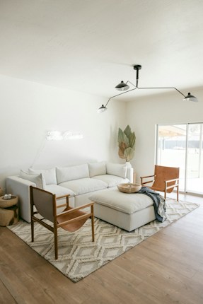 A modern living room featuring a large white sectional sofa with several cushions, accompanied by two leather armchairs with wooden frames. In front of the sofa is a large upholstered ottoman. A soft, patterned area rug covers the wooden floor, and a minimalist, black metal ceiling light fixture hangs above. A potted plant with large leaves is positioned in the corner near a window that allows natural light to flood the room. A small round wooden side table sits next to the sofa with a few items on top.
