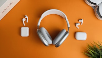 A pair of over-ear headphones along with two pairs of wireless earbuds and their respective charging cases are arranged on an orange surface. A box labeled 'AirPods Max' is partially visible in the top left corner. There is also a small green plant in the bottom right corner.