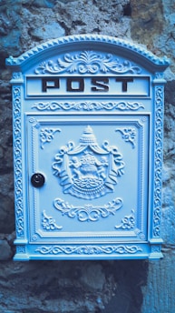 A decorative, ornate mailbox with the word 'POST' embossed at the top. The mailbox is adorned with intricate patterns and designs, including a coat of arms in the center. It is painted in a light pastel blue and is mounted against a textured stone wall, providing a vintage look.