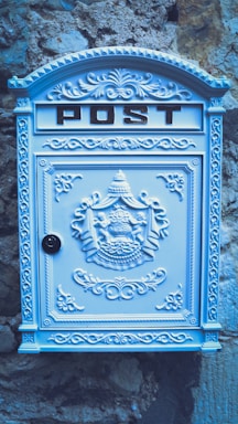 A decorative, ornate mailbox with the word 'POST' embossed at the top. The mailbox is adorned with intricate patterns and designs, including a coat of arms in the center. It is painted in a light pastel blue and is mounted against a textured stone wall, providing a vintage look.