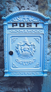 A decorative, ornate mailbox with the word 'POST' embossed at the top. The mailbox is adorned with intricate patterns and designs, including a coat of arms in the center. It is painted in a light pastel blue and is mounted against a textured stone wall, providing a vintage look.