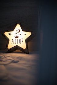 A star-shaped night light softly illuminates a room with a warm glow. The night light features a crescent moon and stars, along with the name 'AITOR' that is prominently displayed in the center. The glow casts gentle shadows on a textured surface beneath.