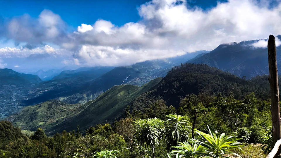 The image depicts a breathtaking view of a mountain range with lush green vegetation and towering trees, set against a clear blue sky dotted with fluffy white clouds. The mountains are blanketed in dense forests, creating a sense of tranquility and harmony between nature and the environment. A small wooden post stands on the right side of the frame, adding an element of human presence to this otherwise untouched natural landscape.