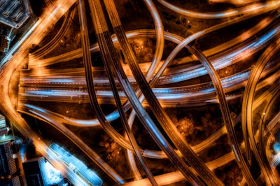 An aerial view of an intricate network of highways and overpasses illuminated by street lights. The lanes crisscross in a complex pattern, with the roads glowing in shades of orange and white from the vehicle lights. The urban landscape surrounding the highways is visible with blocks of buildings and parking lots.