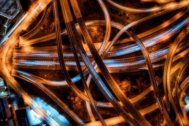 An aerial view of an intricate network of highways and overpasses illuminated by street lights. The lanes crisscross in a complex pattern, with the roads glowing in shades of orange and white from the vehicle lights. The urban landscape surrounding the highways is visible with blocks of buildings and parking lots.