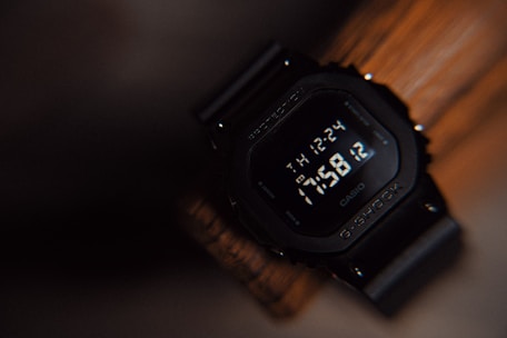 A digital wristwatch with a black case and strap, displaying the time as 17:58:12. The watch features a rugged design with a screen that shows additional information like the day of the week and date. The background is blurred and creates a warm, earthy tone.
