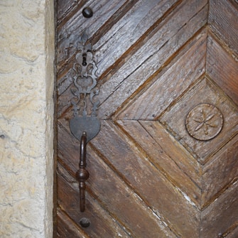 A close-up view of a wooden door with detailed carvings and a prominent metal handle. The wood has a rich, textured appearance with diagonal patterns and an ornate metal lock plate surrounding the keyhole. The door features an additional circular carved design to the right.