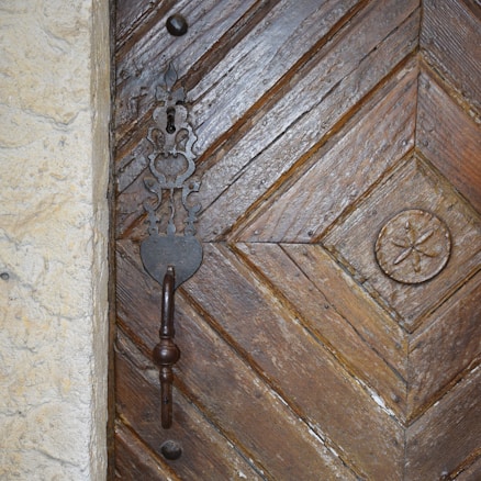 A close-up view of a wooden door with detailed carvings and a prominent metal handle. The wood has a rich, textured appearance with diagonal patterns and an ornate metal lock plate surrounding the keyhole. The door features an additional circular carved design to the right.
