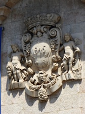 A stone relief sculpture on a wall featuring two cherubs flanking a central shield adorned with the year 1945. The shield is crowned and surrounded by intricate designs including grape clusters and scrollwork. Below the shield is a prominent lion head with more ornamental details.