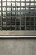 A wall composed of multiple glass blocks arranged in a grid pattern. The blocks are transparent but have a frosted texture, partially obscuring the light and view behind them. Below the glass block section is a horizontal metal rail, with a smooth, reflective surface, and beneath it a section of concrete wall with a slightly rough texture.