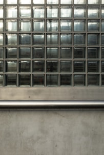 A wall composed of multiple glass blocks arranged in a grid pattern. The blocks are transparent but have a frosted texture, partially obscuring the light and view behind them. Below the glass block section is a horizontal metal rail, with a smooth, reflective surface, and beneath it a section of concrete wall with a slightly rough texture.