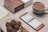 A neatly arranged table setting with a cup of hot chocolate next to a smartphone displaying a menu in a foreign language. Nearby, a plate holds several chocolate cookies alongside a small wooden fork. A partially visible book and a boxed product labeled 'smartisan' are also present.