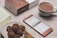 A neatly arranged table setting with a cup of hot chocolate next to a smartphone displaying a menu in a foreign language. Nearby, a plate holds several chocolate cookies alongside a small wooden fork. A partially visible book and a boxed product labeled 'smartisan' are also present.