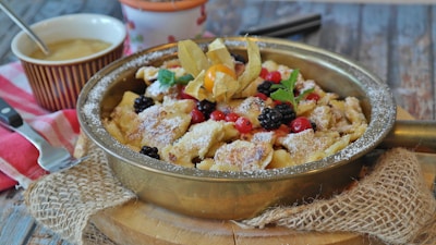 A rustic pan filled with a traditional dessert topped with powdered sugar and assorted berries, such as blackberries and red currants. Garnished with a sprig of mint and a decorative husk cherry. The setting includes a wooden surface, a checkered napkin, and a small bowl of sauce with a spoon beside it.
