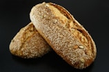 Two loaves of bread encrusted with sesame seeds and other seeds are placed together on a dark surface. The bread has a golden-brown crust and appears freshly baked.