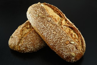 Two loaves of bread encrusted with sesame seeds and other seeds are placed together on a dark surface. The bread has a golden-brown crust and appears freshly baked.
