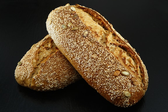 Two loaves of bread encrusted with sesame seeds and other seeds are placed together on a dark surface. The bread has a golden-brown crust and appears freshly baked.