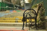 A series of colorful steps leading upward in an outdoor setting, bordered by metal railings and a green fence. A unique metal bench with circular patterns is positioned in the foreground, set against a backdrop of trees and foliage.