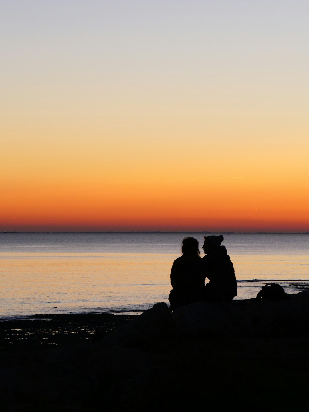 silhouette of 2 people sitting on rock near sea during sunset