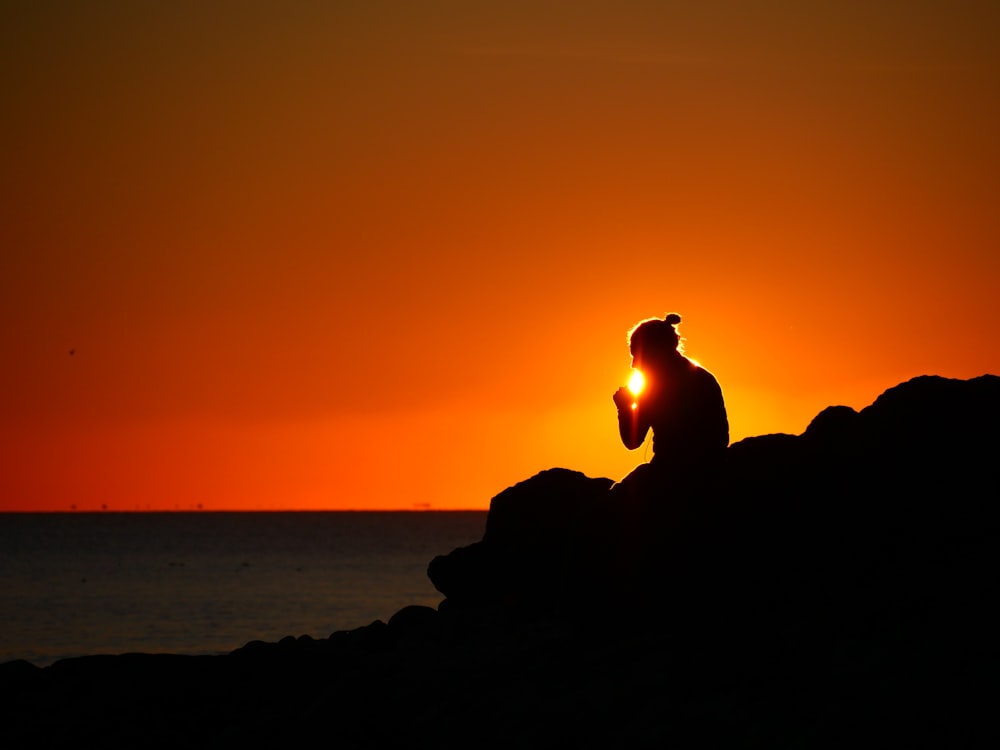 silhouette of person sitting on rock near body of water during sunset