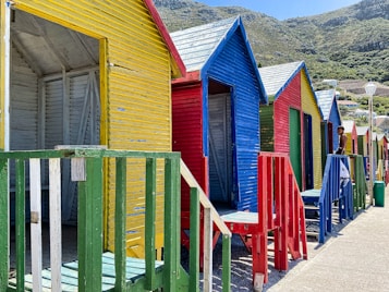 A row of colorful beach huts with bright yellow, blue, red, and green paint. The wooden structures are lined along the beachside, set against a backdrop of lush, green hillside. A person is standing on the red wooden steps of one of the cabins.
