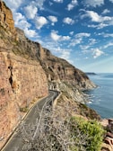 gray asphalt road beside brown rocky mountain beside body of water during daytime