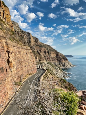 gray asphalt road beside brown rocky mountain beside body of water during daytime