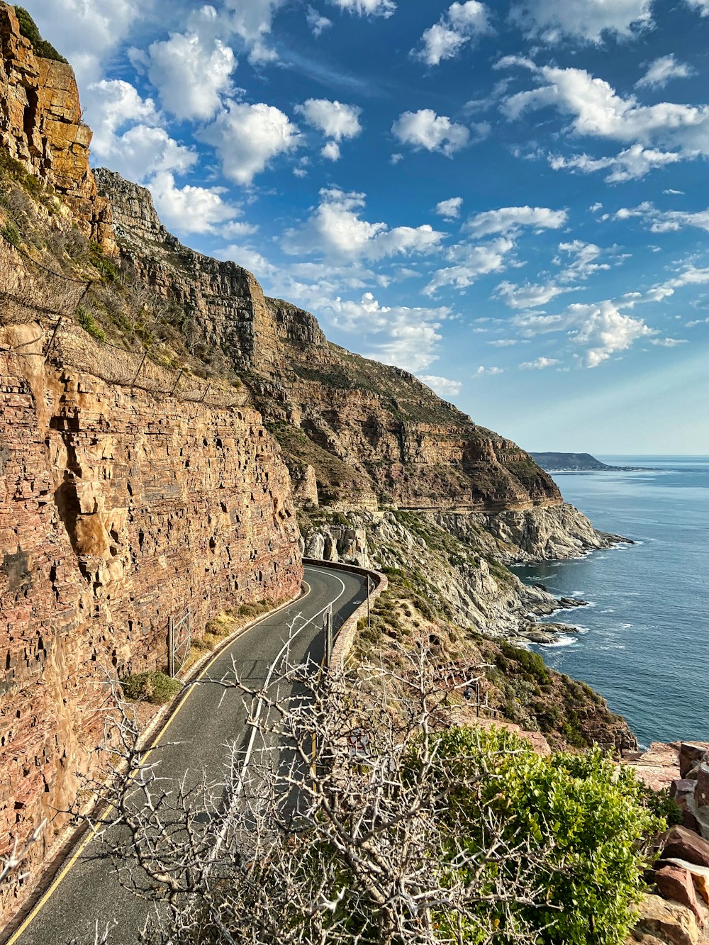 gray asphalt road beside brown rocky mountain beside body of water during daytime