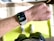 A close-up view of a person's wrist wearing a smartwatch displaying a workout mode for High Intensity Interval Training. The background includes a bright green exercise mat on a wooden floor. The person is wearing white athletic shoes and dark shorts.