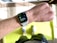 A close-up view of a person's wrist wearing a smartwatch displaying a workout mode for High Intensity Interval Training. The background includes a bright green exercise mat on a wooden floor. The person is wearing white athletic shoes and dark shorts.