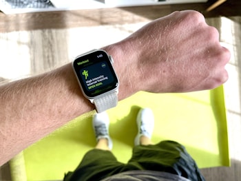 A close-up view of a person's wrist wearing a smartwatch displaying a workout mode for High Intensity Interval Training. The background includes a bright green exercise mat on a wooden floor. The person is wearing white athletic shoes and dark shorts.