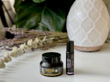 A small black container labeled CBD Muscle Rub and a black roller bottle labeled Calm are placed on a white surface. Behind them, there is a white, patterned diffuser. There are also dried lavender and eucalyptus stems arranged on the left side. In the background, large green leaves provide a soothing backdrop.