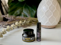 A small black container labeled CBD Muscle Rub and a black roller bottle labeled Calm are placed on a white surface. Behind them, there is a white, patterned diffuser. There are also dried lavender and eucalyptus stems arranged on the left side. In the background, large green leaves provide a soothing backdrop.