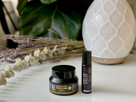A small black container labeled CBD Muscle Rub and a black roller bottle labeled Calm are placed on a white surface. Behind them, there is a white, patterned diffuser. There are also dried lavender and eucalyptus stems arranged on the left side. In the background, large green leaves provide a soothing backdrop.