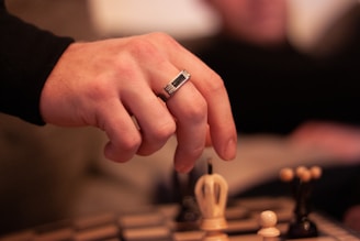 A person's hand adorned with a ring is making a move on a chessboard. The focus is on the hand and the chessboard, creating a sense of concentration and strategy. The background is blurred, suggesting an indoor, intimate setting.
