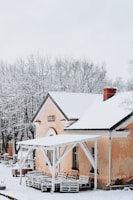 A rustic building with snow-covered rooftops and surrounding trees. The structure has a warm, earthy tone with a sign that reads 'Ergli'. Wooden pallets are stacked under the canopy, indicating a rural or quaint setting.