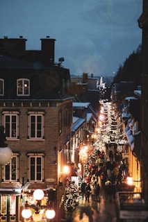 A charming, narrow street bustling with people enjoying festive holiday lights and decorations. Buildings are adorned with glowing ornaments and strings of lights, creating a warm and inviting atmosphere in an evening setting. The street is framed by architecturally rich buildings, and there is a visible Christmas tree decorated with lights.