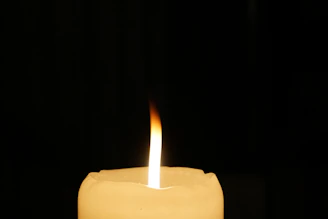 A single candle flame burns with a gentle flicker, casting a warm glow against a dark background. The candle wax is smooth and creamy, with the wick standing upright at the center of the flame.