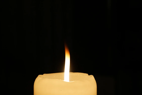 A single candle flame burns with a gentle flicker, casting a warm glow against a dark background. The candle wax is smooth and creamy, with the wick standing upright at the center of the flame.