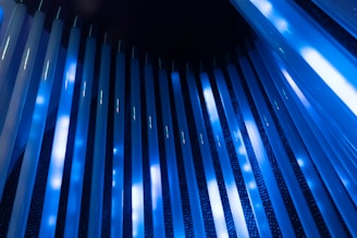 Vertical rows of glowing blue and white lights arranged in a cylindrical pattern create a futuristic and abstract visual. The light is reflected off what appears to be a metallic or glossy surface, enhancing the overall brightness and contrast.
