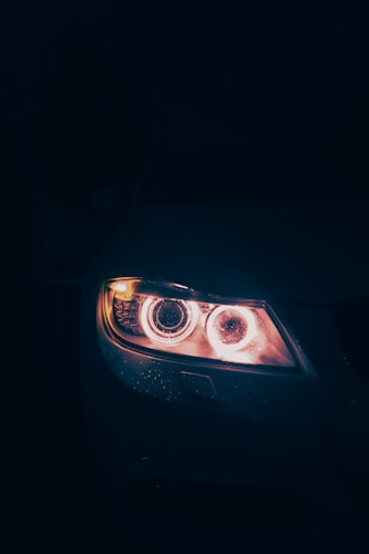 A close-up of a car headlight illuminated in the dark. The light casts a warm, intense glow and the surface around it displays water droplets, indicating recent rain or moisture. The background is dark, creating a dramatic contrast.